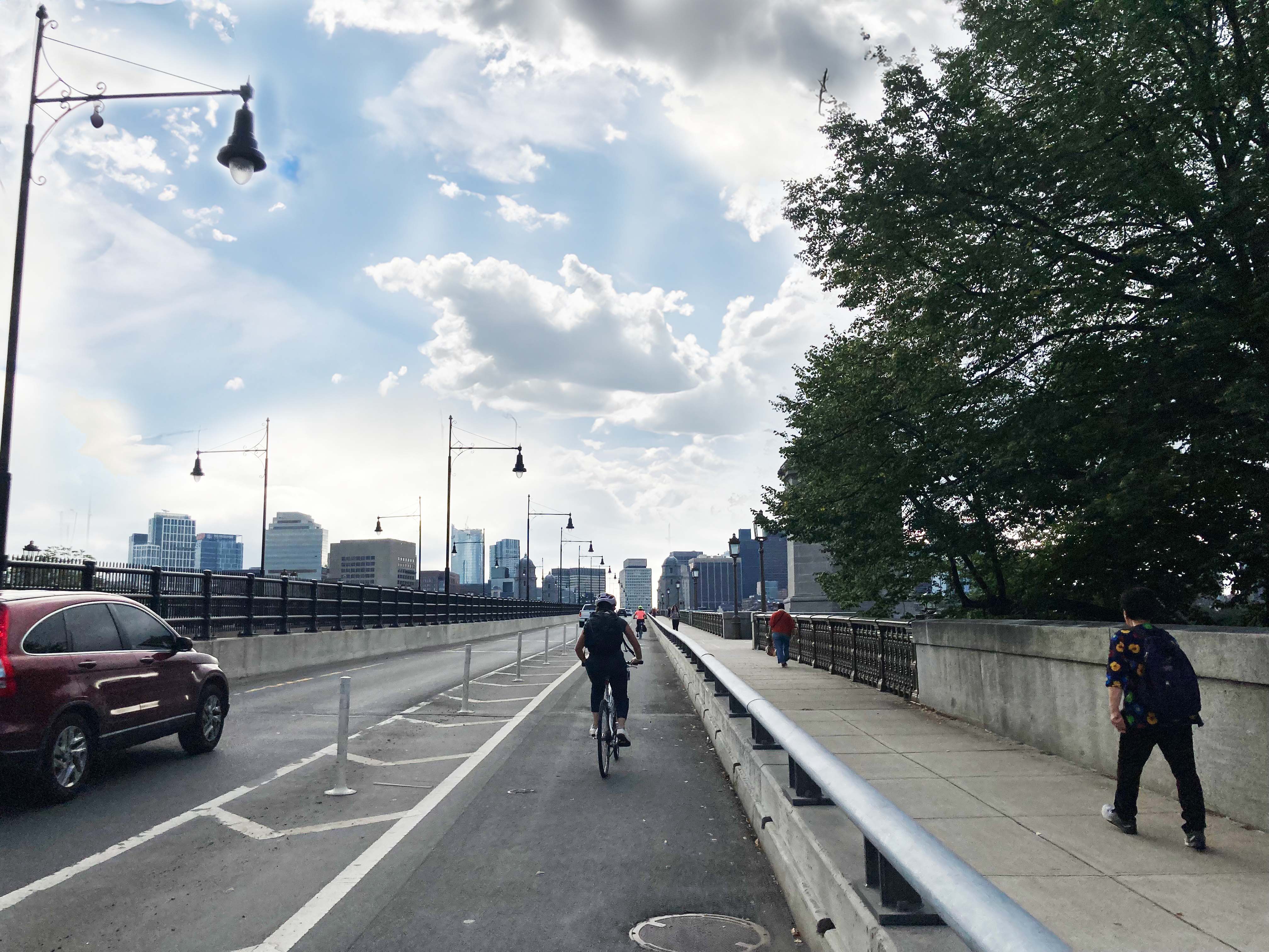 Road shared by pedestrians, cyclists, and a car with a city skyline in the background. Image Credit: FHWA