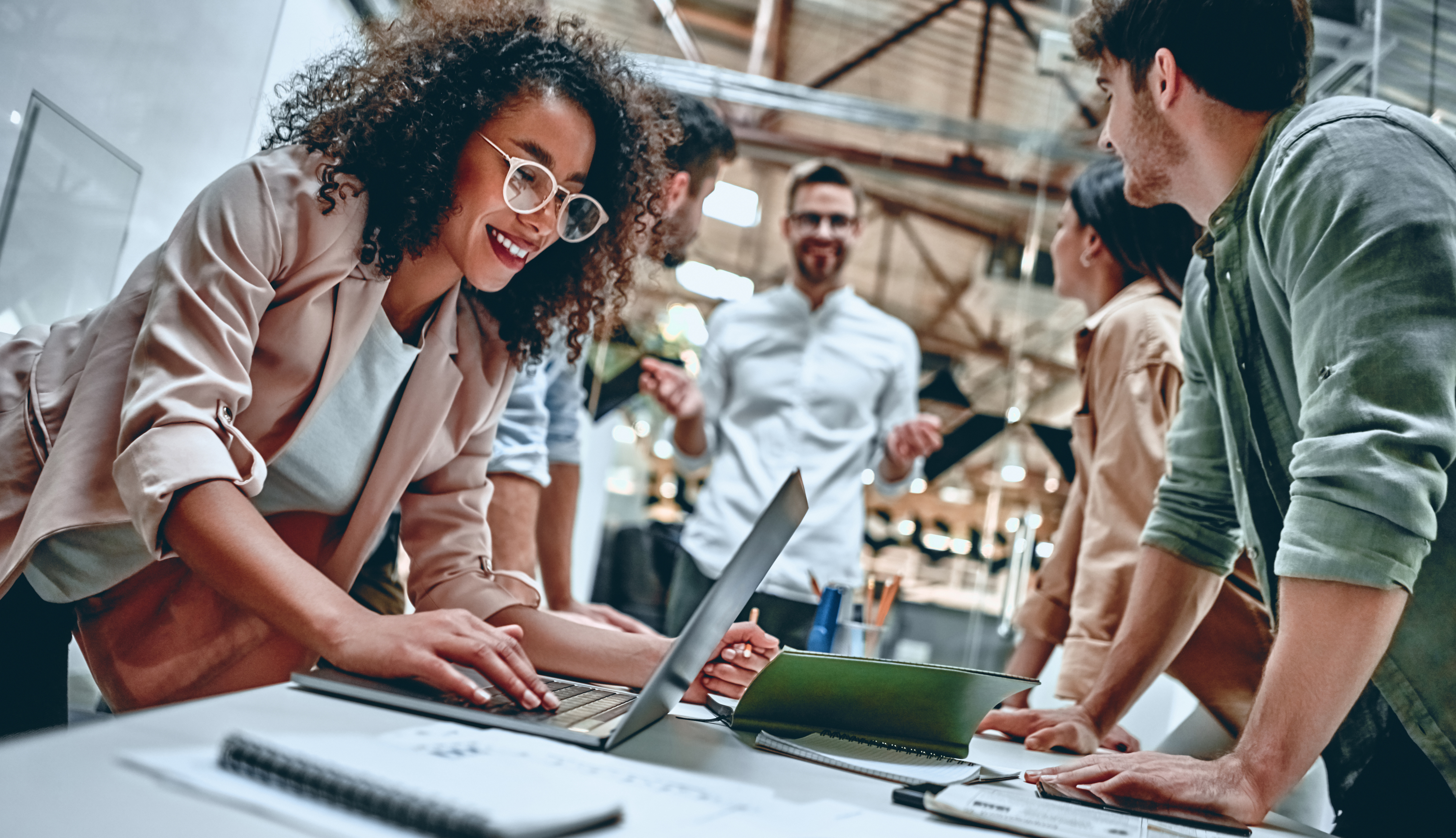 woman smiling and standing over her laptop with coworkers around a table. Image Credit: AdobeStock_300144034