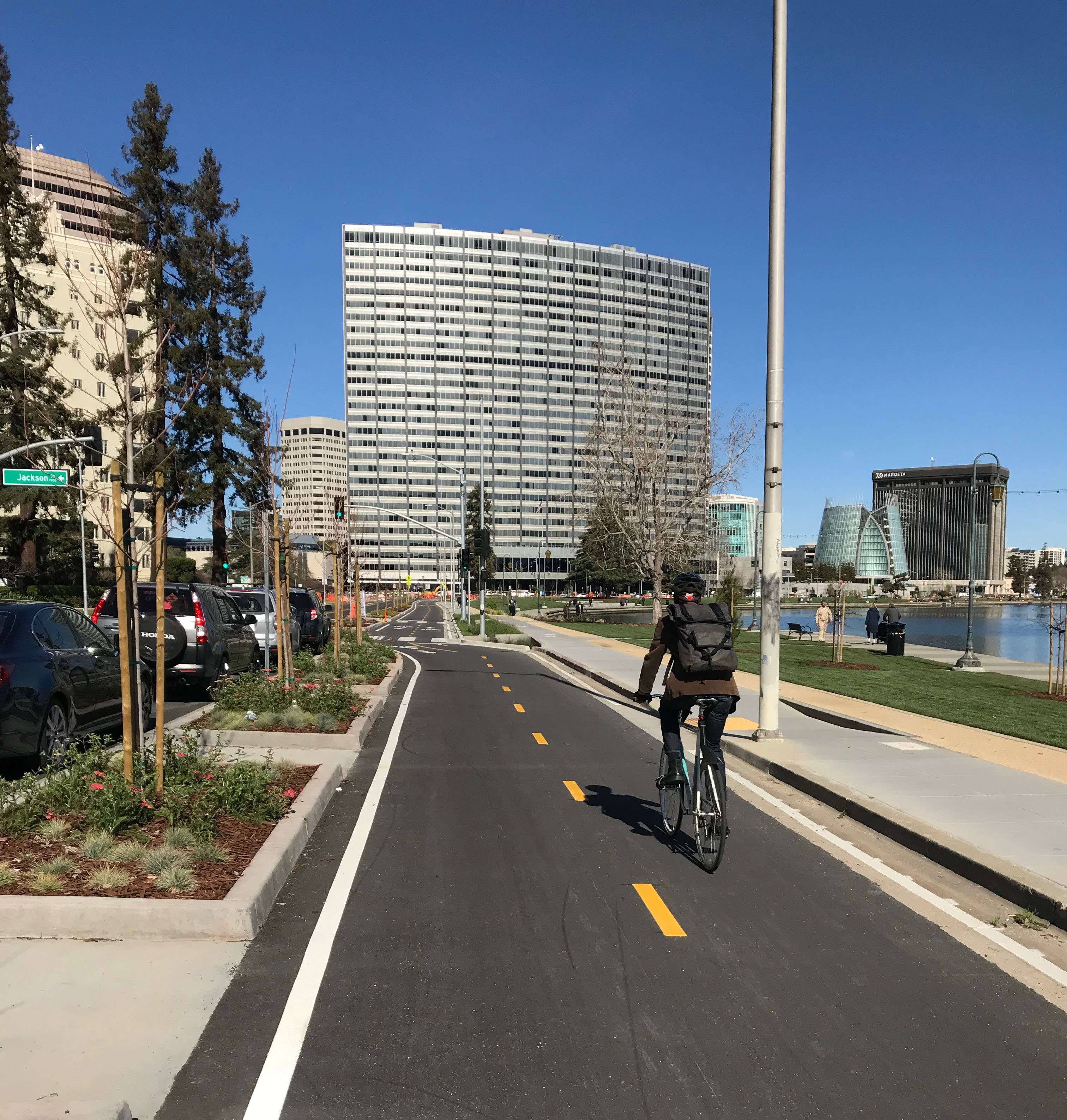 Cyclist on bike path alongside a road with traffic, with tall city buildings and a blue sky in the background. Image Credit: FHWA