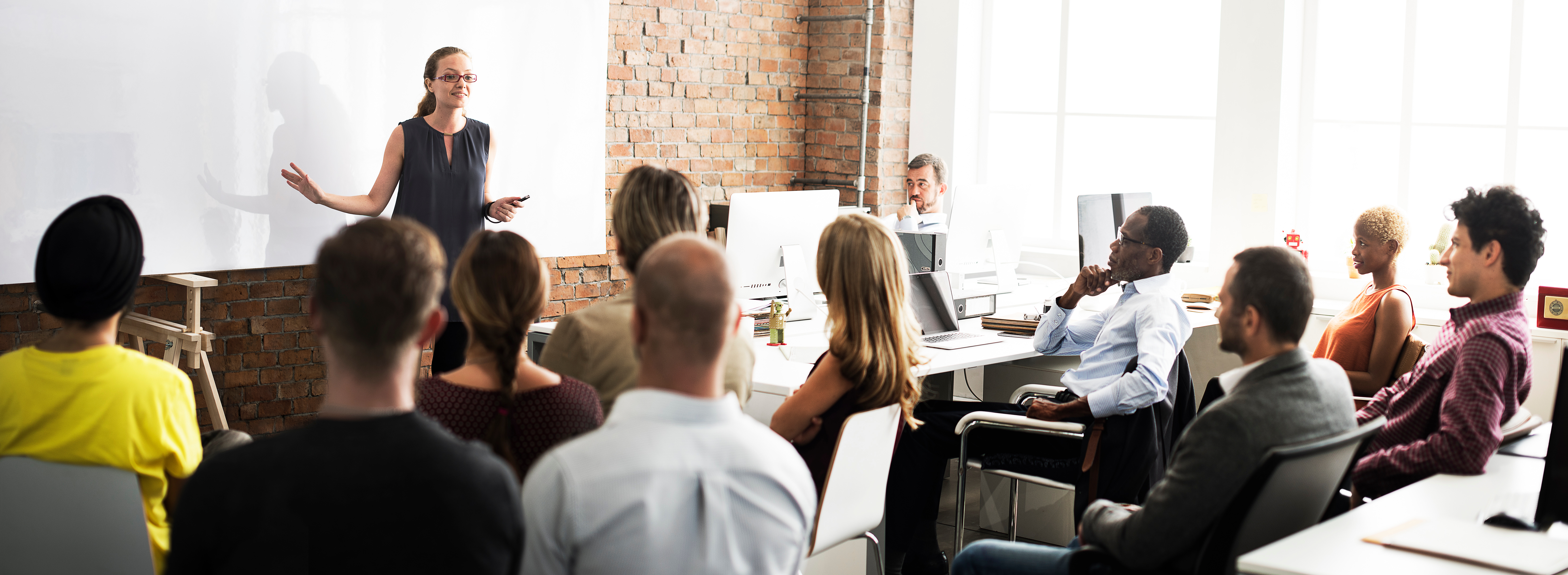 woman teaching in front of a class of people sitting at desks in an office. Image Credit: AdobeStock_105568884