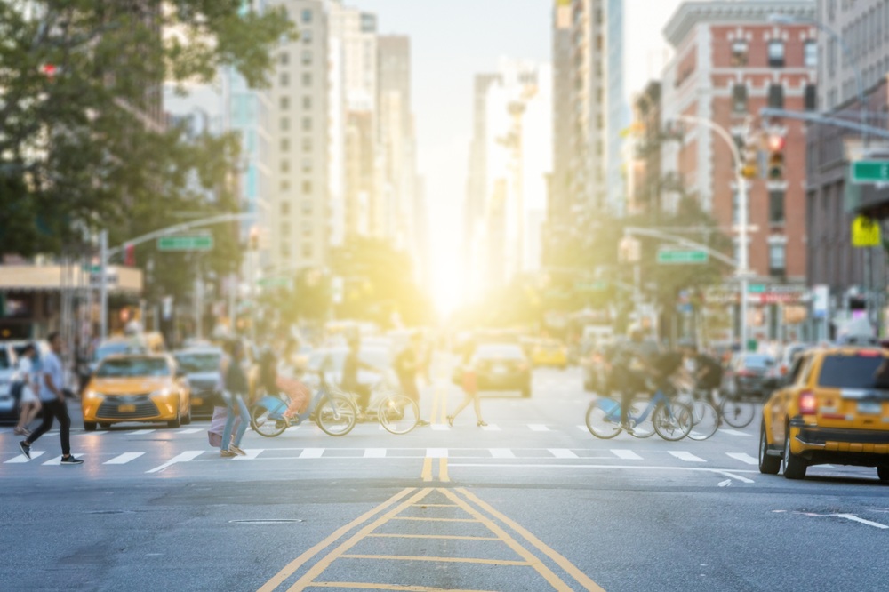 busy crosswalk with pedestrians and bikers crossing in front of traffic, bright sun between streets lined with tall city buildings. Image Credit: AdobeStock_172277527